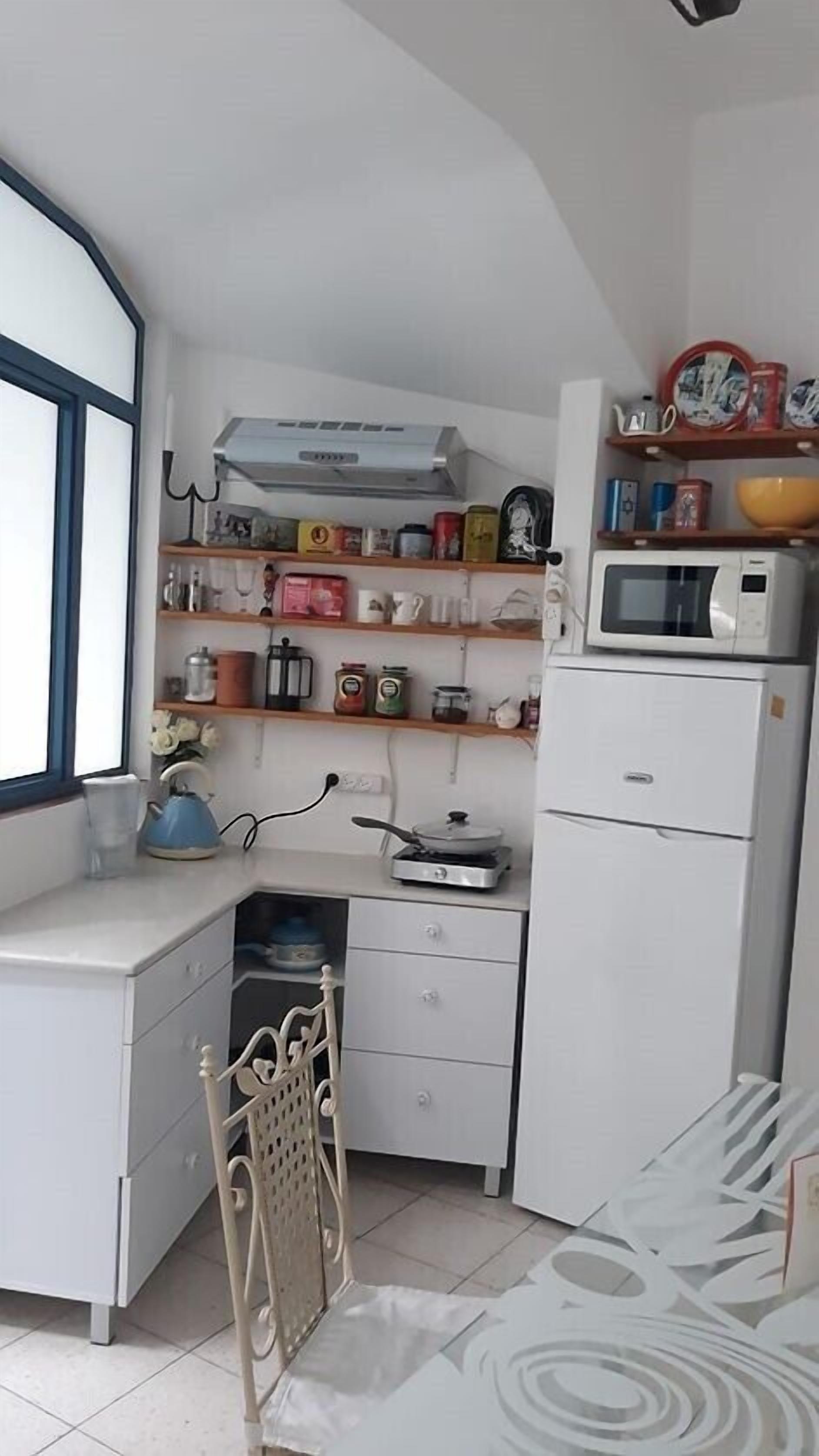 Cocina compacta con armarios blancos, microondas, hervidor y varios utensilios y electrodomésticos en estantes.