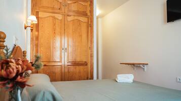 Una habitación con un armario de madera, una cama con edredón verde, una mesita de noche pequeña con una lámpara y un televisor montado en la pared.