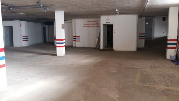 An empty parking garage with white pillars marked with red and blue stripes, a concrete floor, and a door leading to another area. - Hotel Valmar (Villar de Olalla)