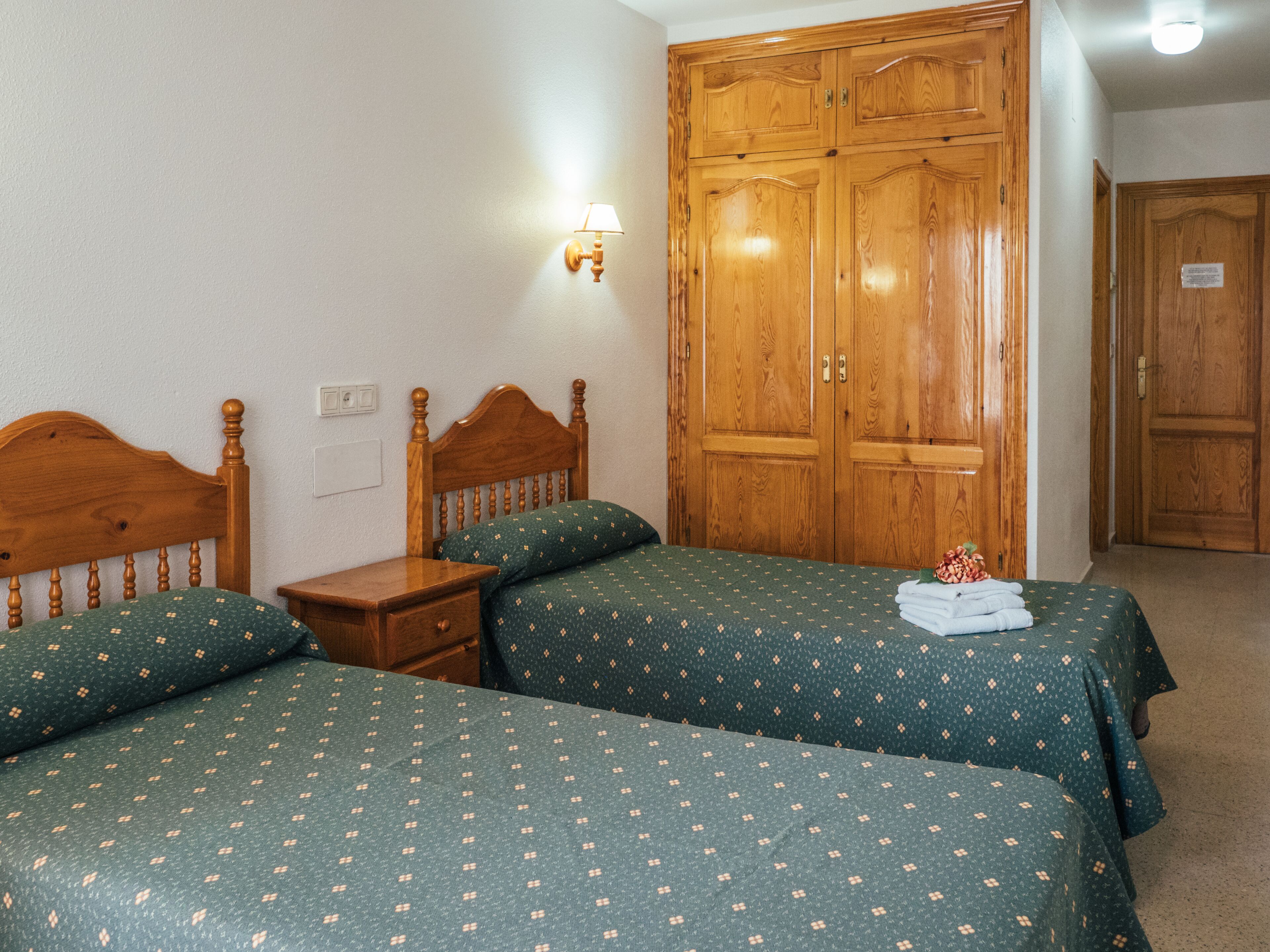 Un dormitorio con dos camas de madera, una mesita de noche, una lámpara de pared y un armario de madera.