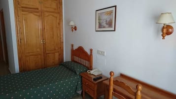 A bedroom with a wooden wardrobe, a bed with a green patterned bedspread, a bedside table with a lamp, and a wall-mounted light fixture.