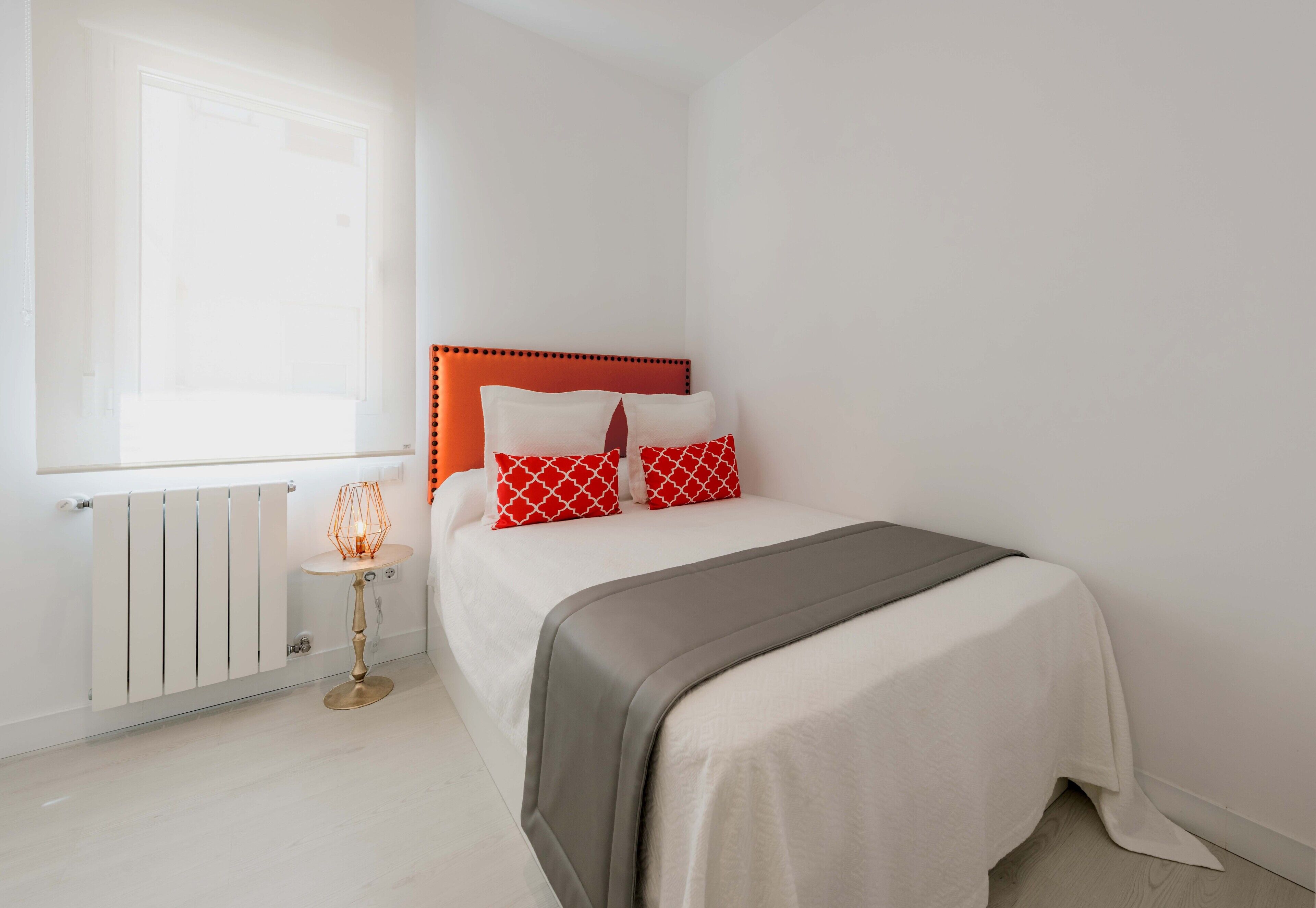 A bedroom with a bed, a radiator, a small table with a lamp, and a window.