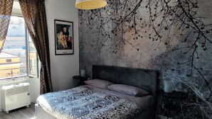 Ein Schlafzimmer mit Bett, Nachttisch, Lampe, Wandgemälde und einem Fenster mit Vorhängen.
