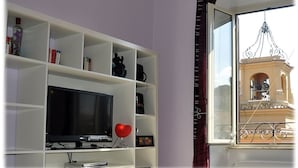 Ein Wohnzimmer mit Fernseher, Regalen mit Büchern und Dekorationsobjekten sowie Blick auf ein Gebäude durch ein Fenster.