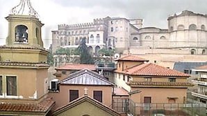 Blick auf ein historisches Gebäude mit Glockenturm und Kuppel, umgeben von weiteren Gebäuden mit roten Ziegeldächern.