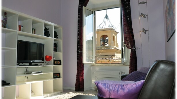 Ein Wohnzimmer mit Fernseher auf einem weißen Regal, einem Fenster mit Blick auf ein Gebäude und einem schwarzen Sessel mit einem violetten Kissen.
