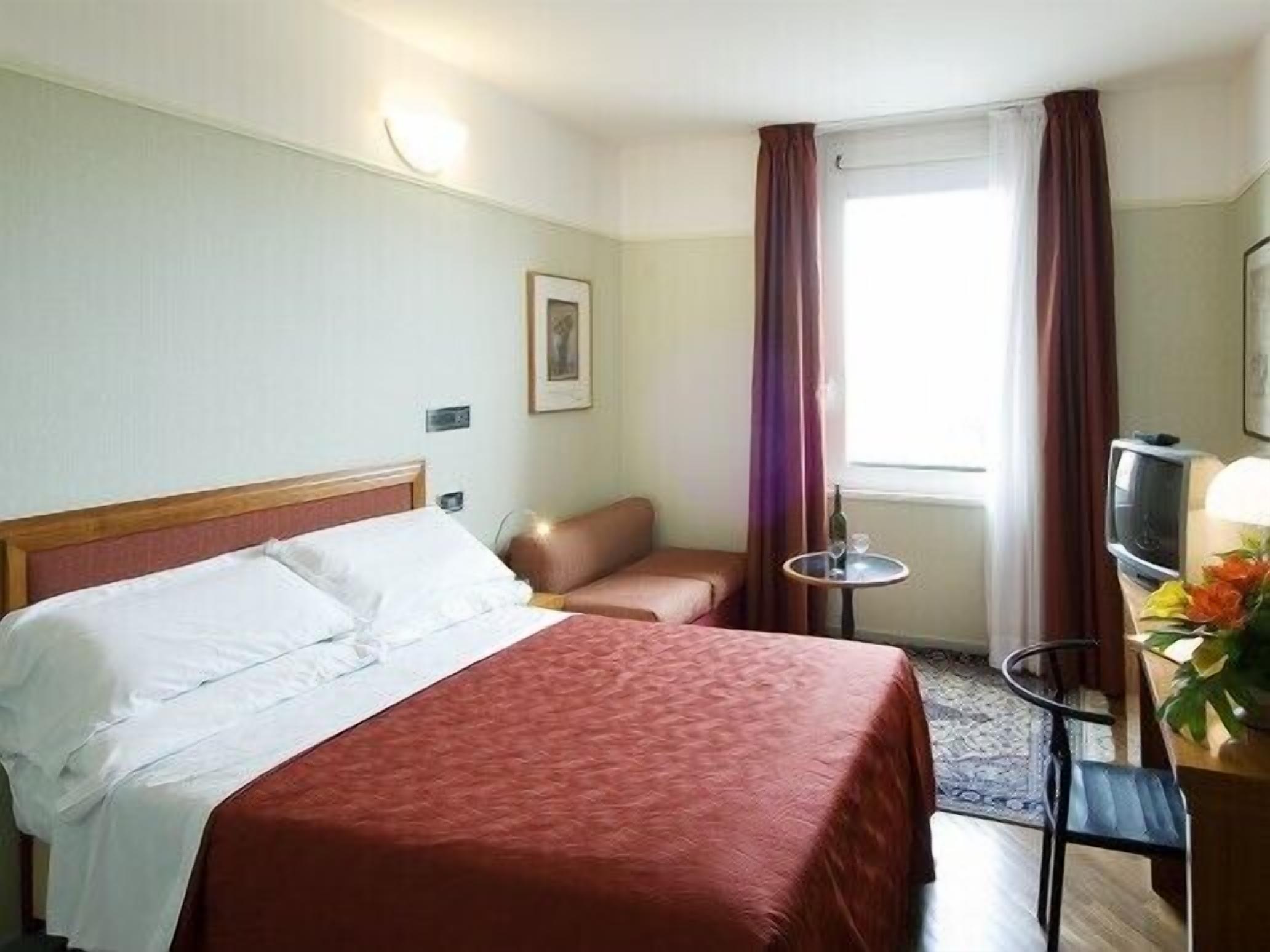 Habitación de hotel con cama, una silla, una mesita con una botella y vaso, y un televisor.