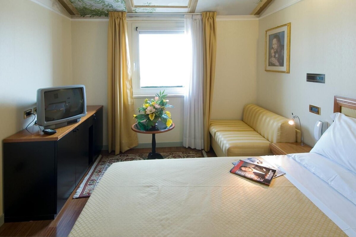 A hotel room with a bed, a television on a desk, a small table with a vase of flowers, and a window with curtains.