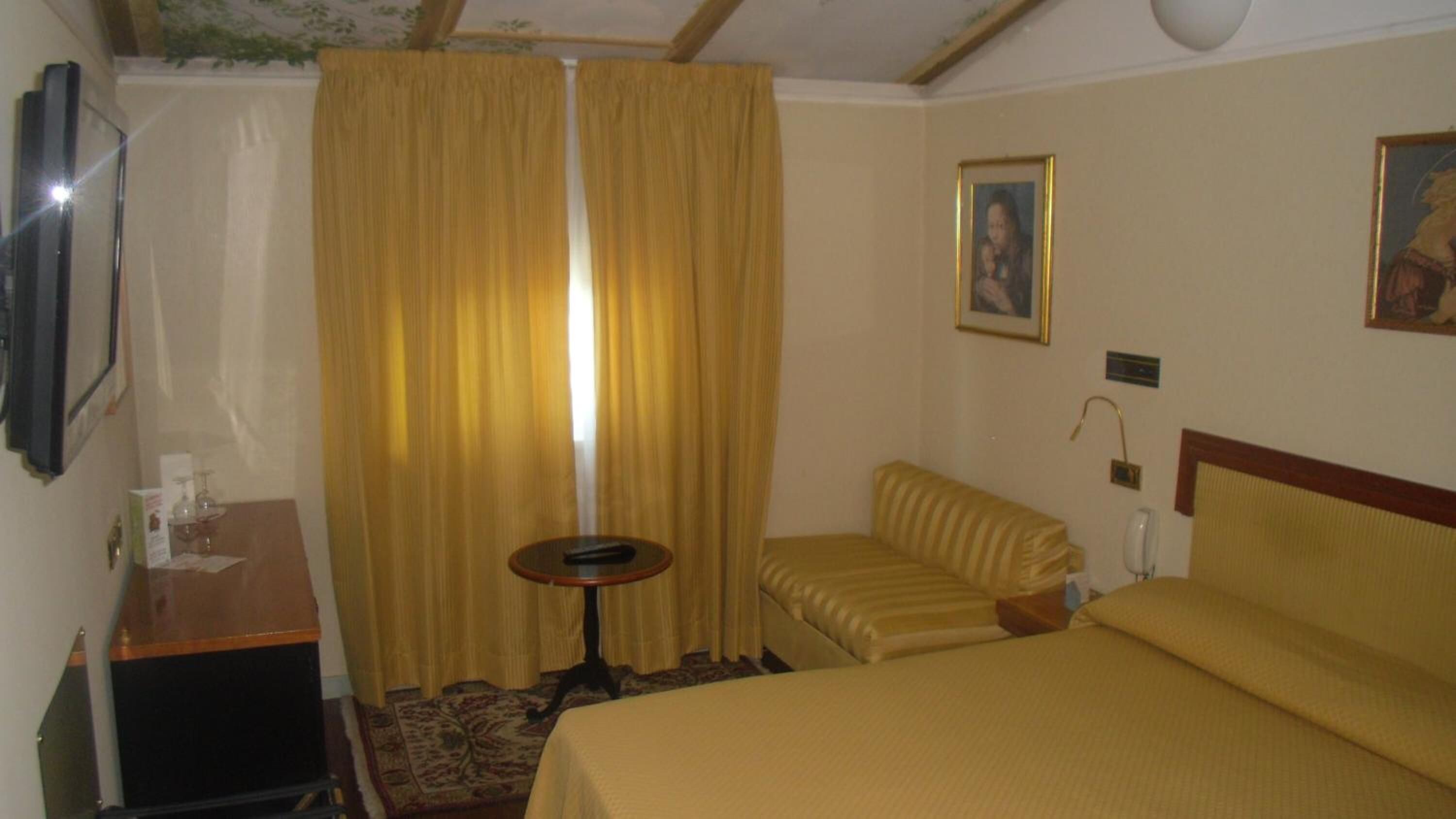 Hotellihuone, jossa on sänky, pieni pöytä, tuoli, työpöytä, verho ja taulu seinällä.