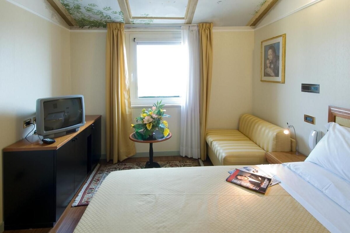 A hotel room with a bed, a TV on a wooden cabinet, a small table with flowers, a sofa, and a picture on the wall.