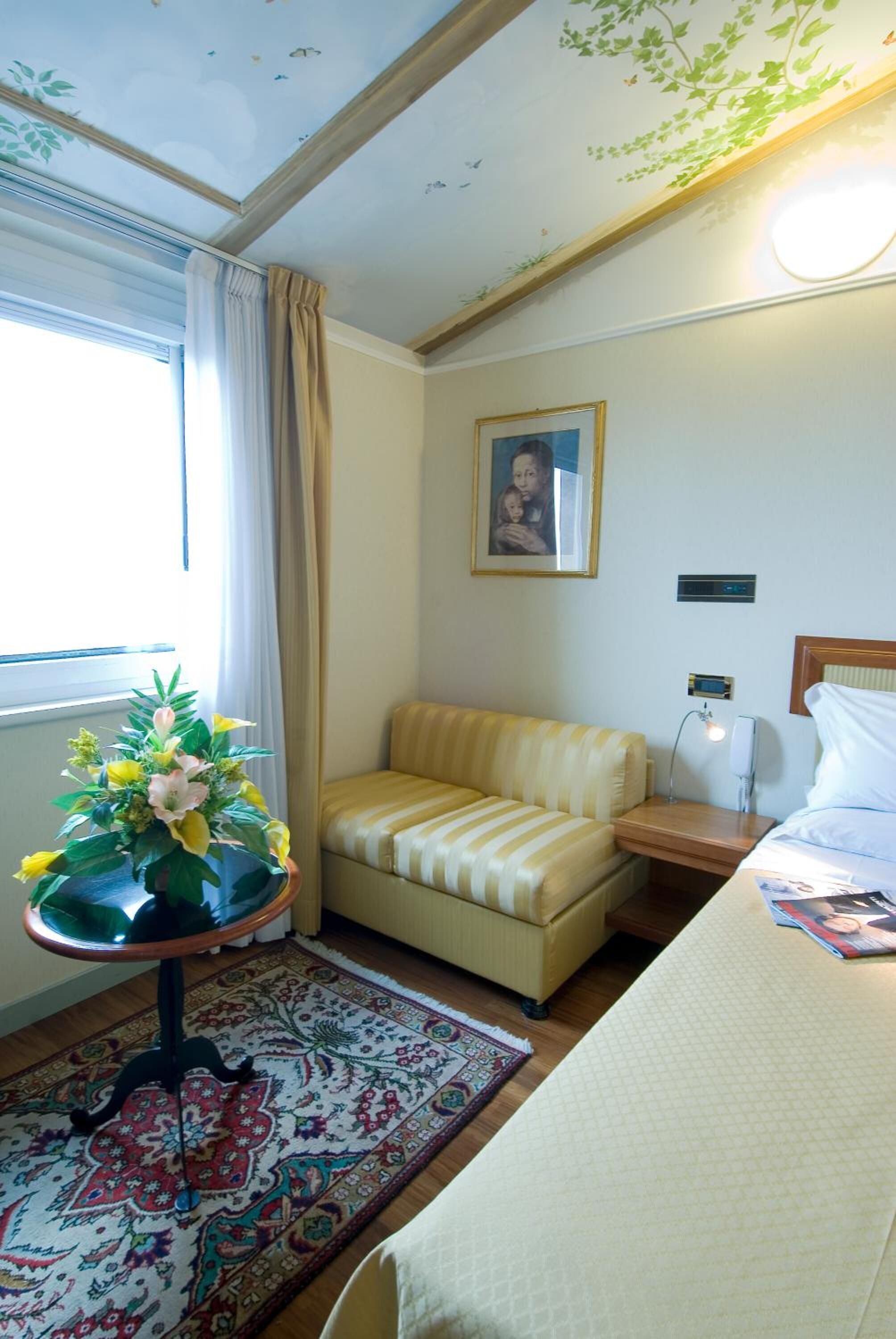 Habitación de hotel con cama, un pequeño sofá, una mesa con un florero, una mesita de noche, una lámpara y un cuadro en la pared.