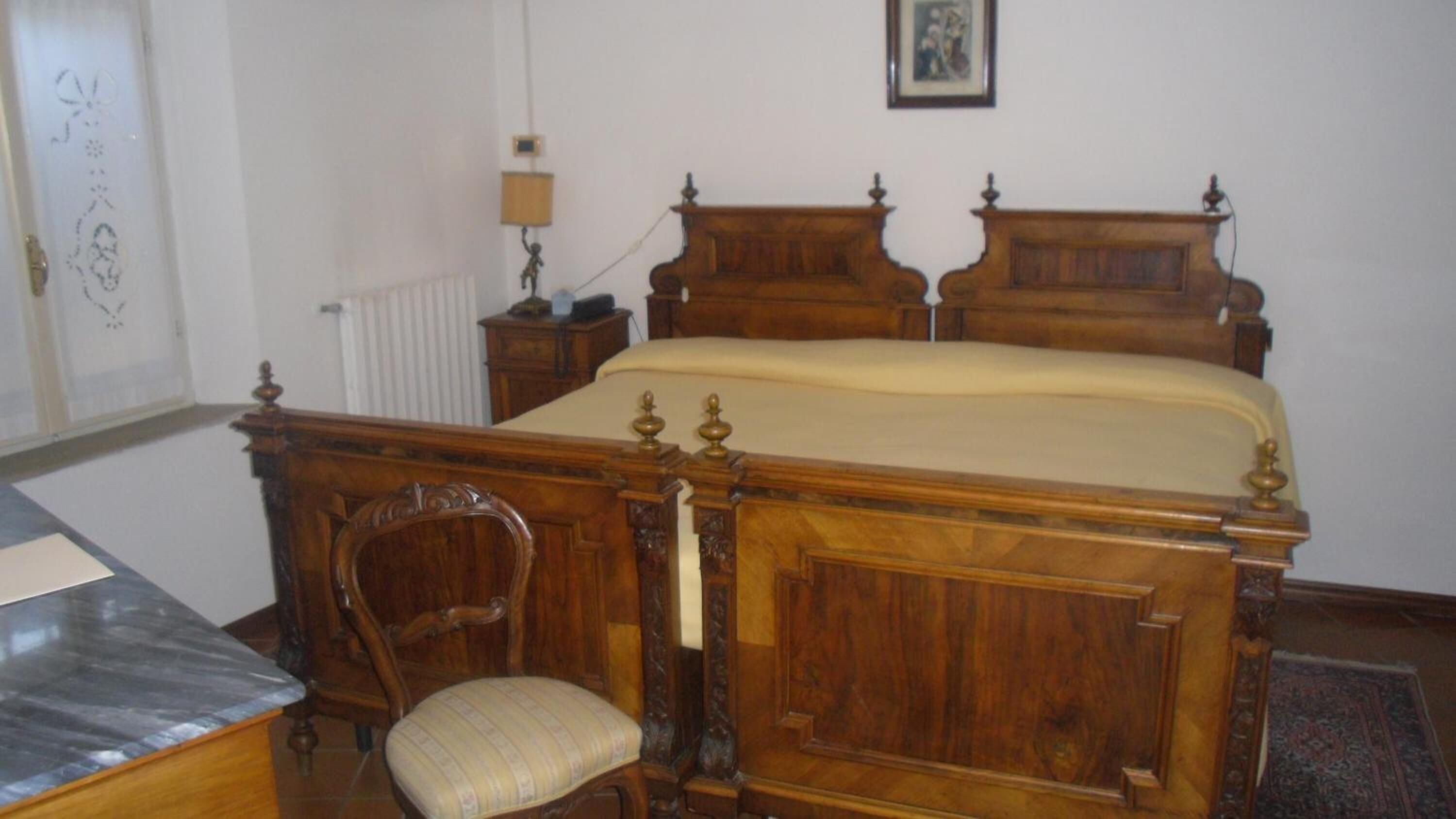 Una cama de madera con cabecera, una mesita de noche, una lámpara y una silla en una habitación con paredes blancas.