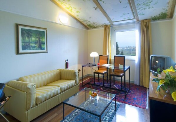 Habitación de hotel con sofá amarillo, una mesa de centro de cristal, una mesa de comedor con sillas, un televisor y un cuadro en la pared.