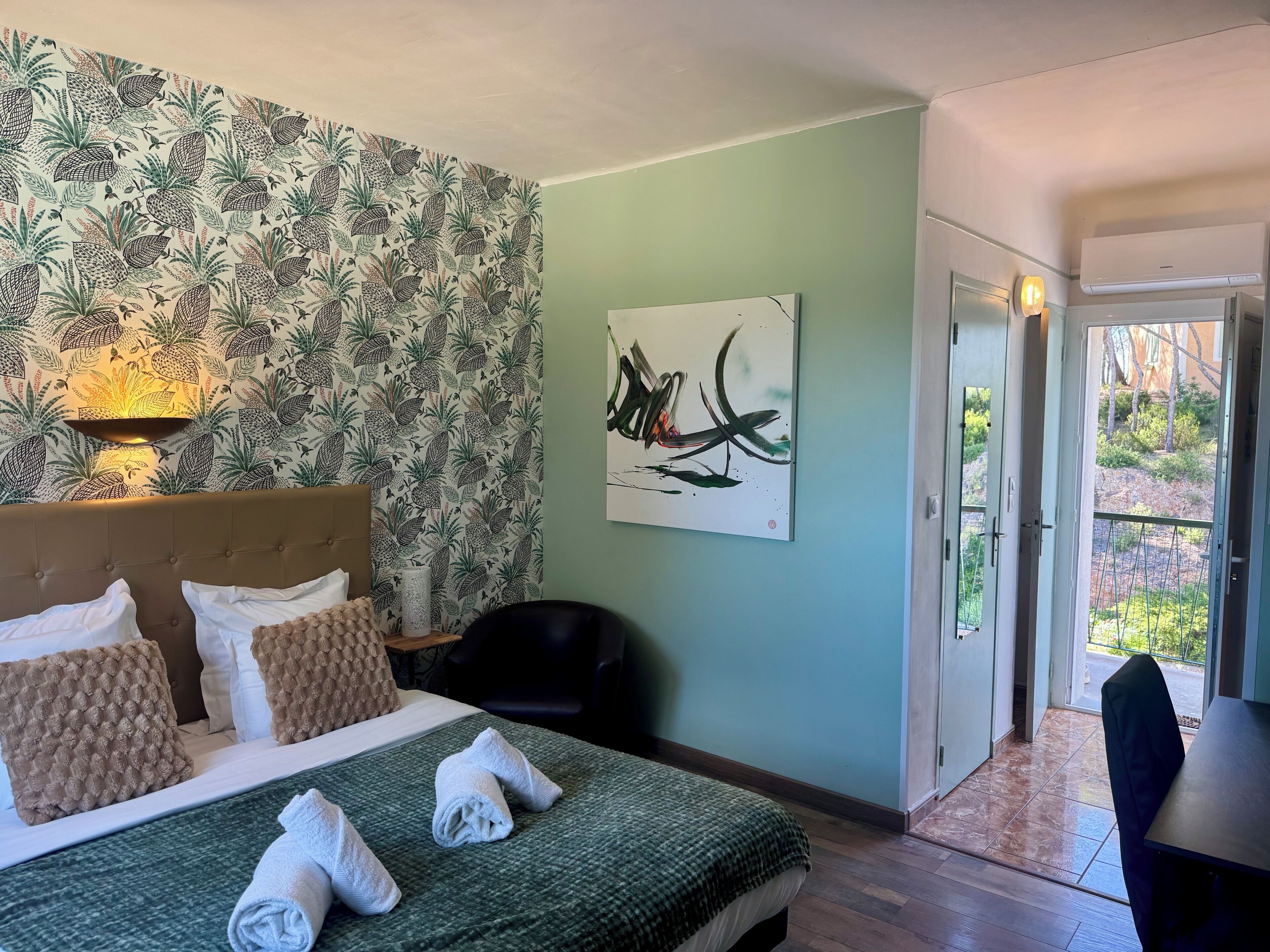 Habitación de hotel con cama, una obra de arte en la pared, una puerta que da a un balcón y una mesita de noche con lámpara.