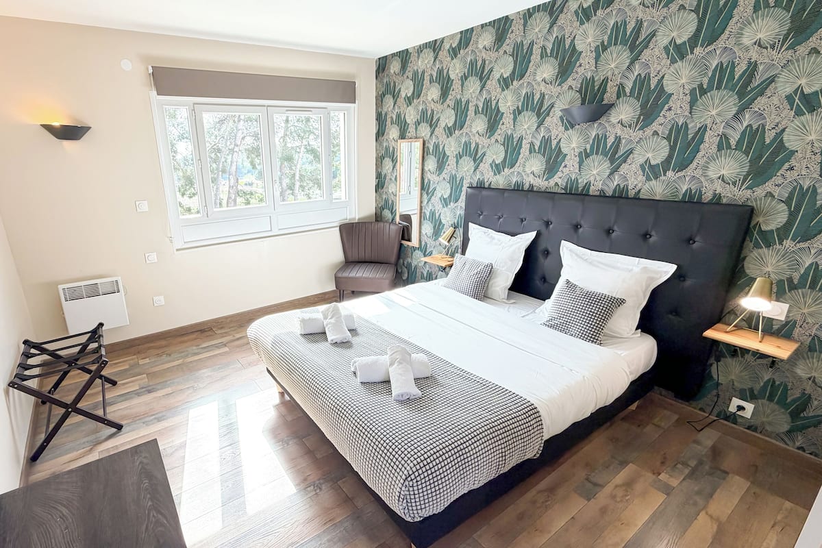 A modern hotel room with a large bed, a window with a view, a wall-mounted TV, and a power outlet.