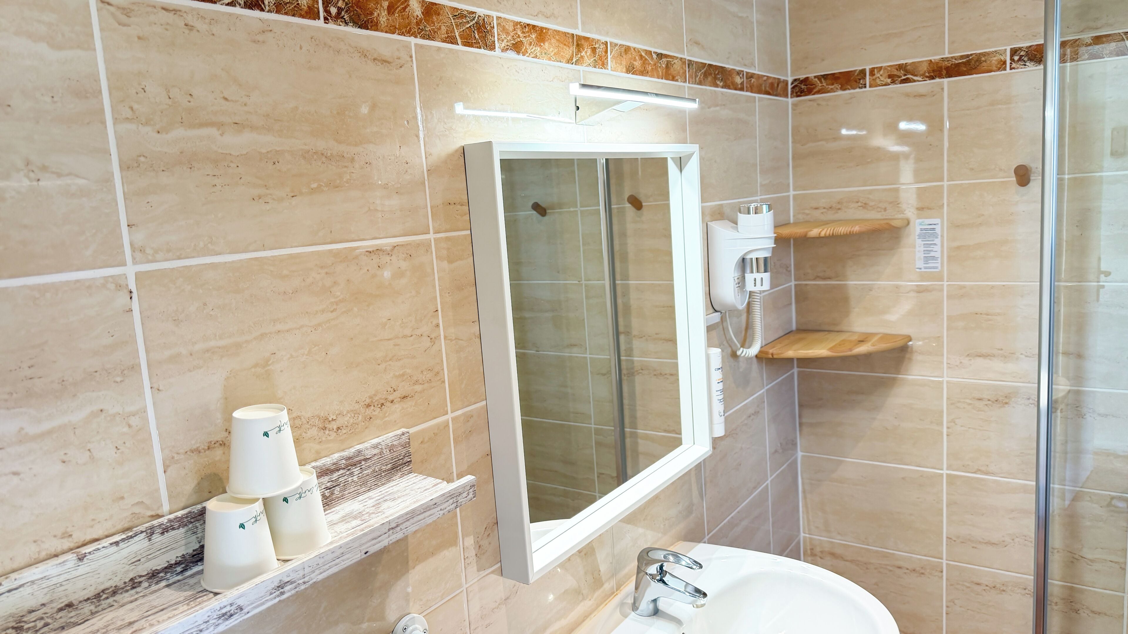A bathroom with a white sink, a mirror, a glass shower enclosure, and a wooden shelf with toiletries.