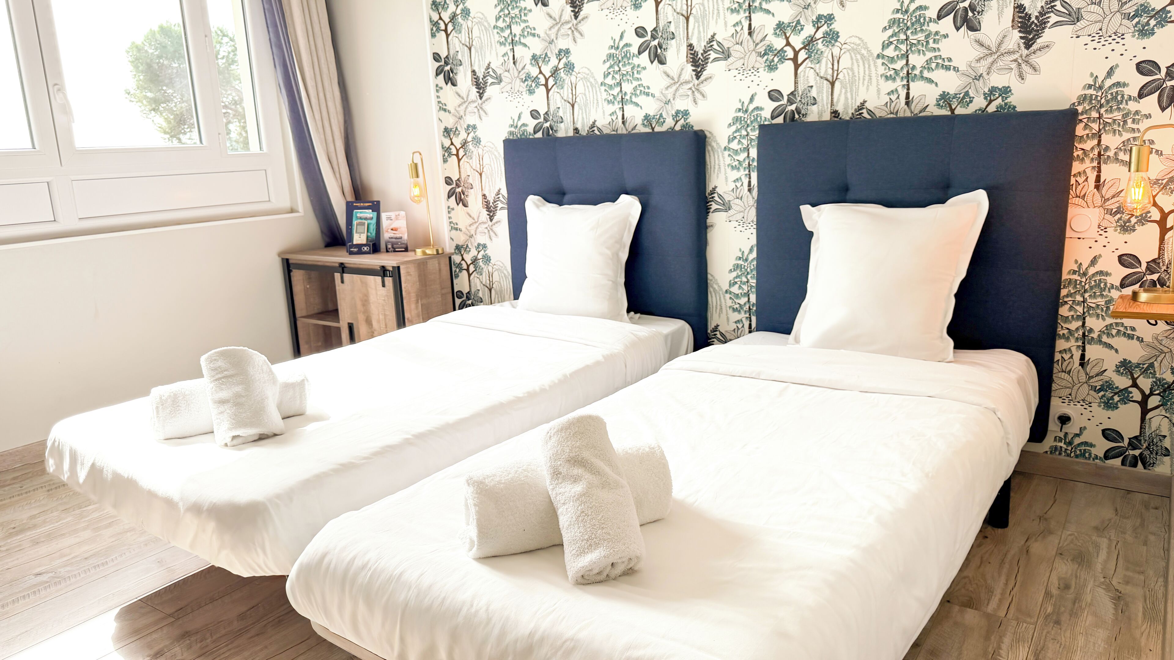 Two single beds with white linens and towels, a small wooden side table with a lamp, and a floral wallpaper pattern.