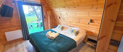 Un dormitorio de cabaña de madera con una cama, una mesita de noche pequeña y una ventana que da a un jardín.