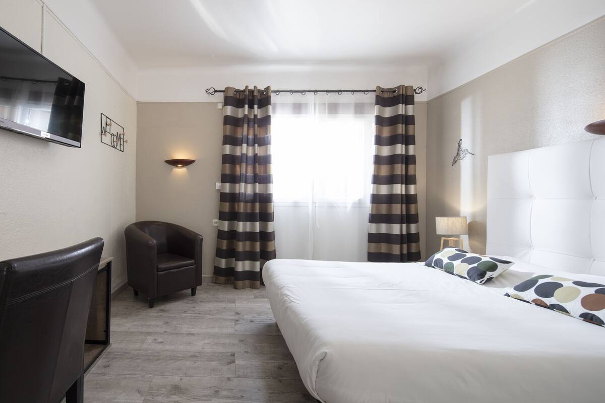 A hotel room with a bed, a chair, a TV, curtains, and a wall-mounted lamp.