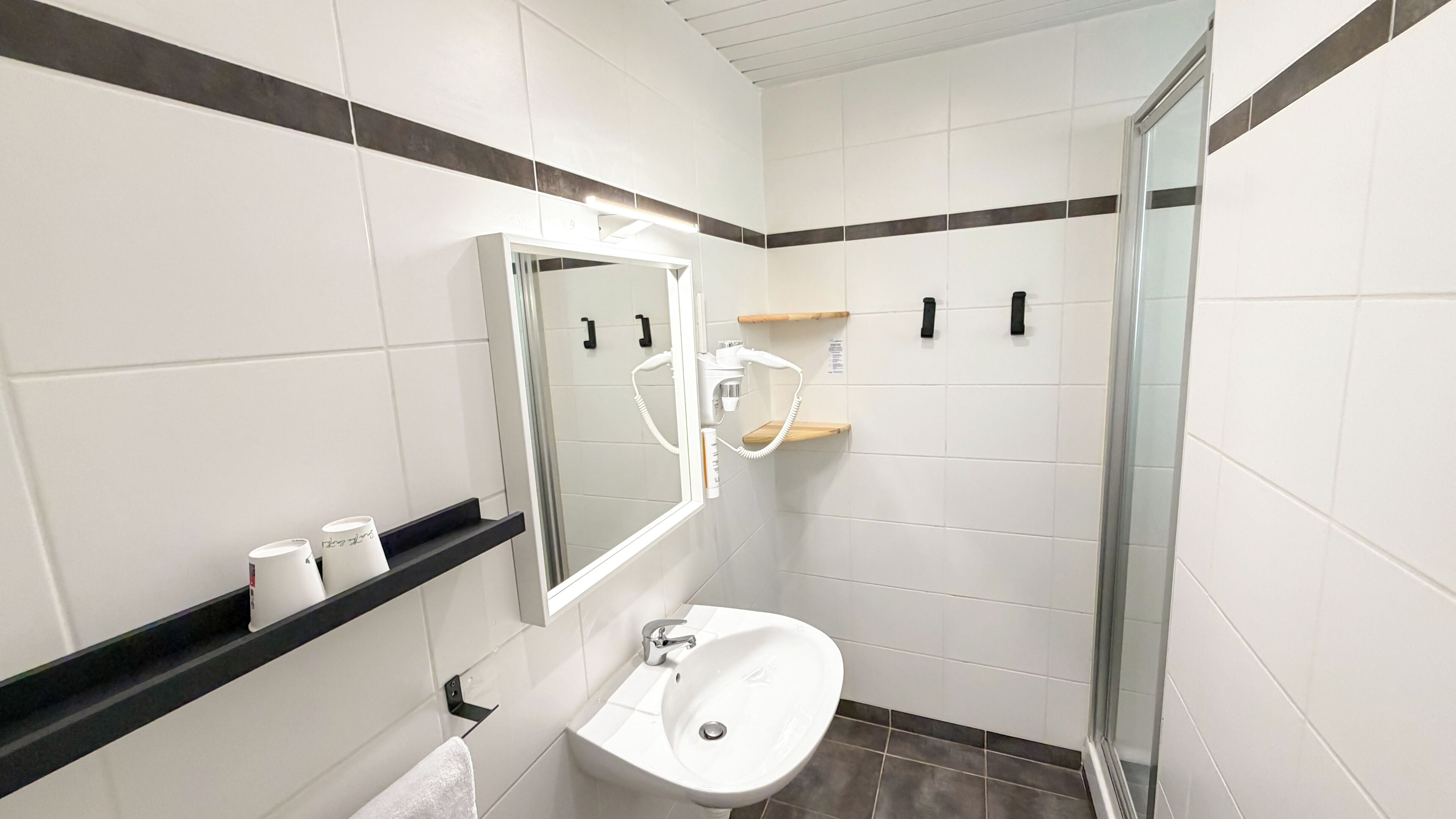 A modern bathroom with a white sink, mirror, and shower. There are toiletries on the shelf and a towel hanging on the wall.
