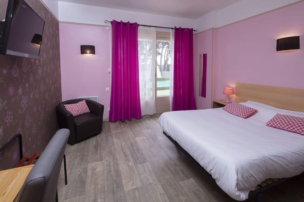 A bedroom with a bed, a chair, a TV, and a window with curtains.