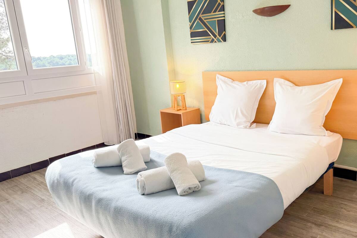 A neatly made bed with white linens and towels, two bedside lamps, and a view of the outdoors through a window.