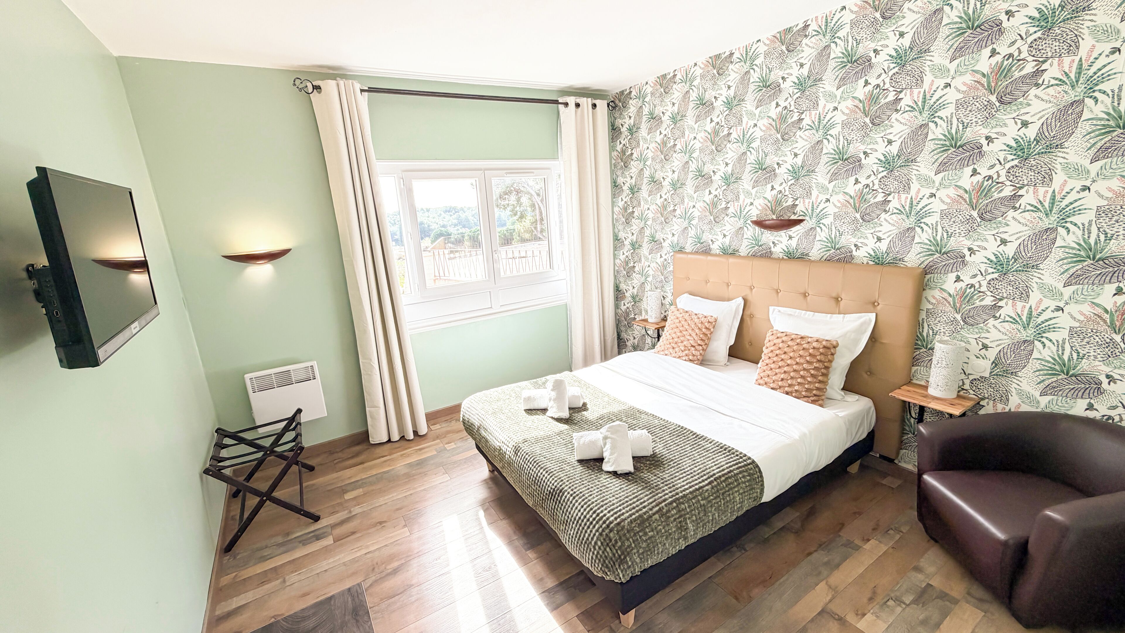 Quarto bem arrumado com cama, poltrona, televisão e janela com cortinas.