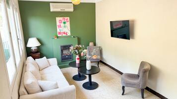 Un salon avec un mur vert, une cheminée, un canapé, un fauteuil, une petite table ornée de vases et un téléviseur fixé au mur.