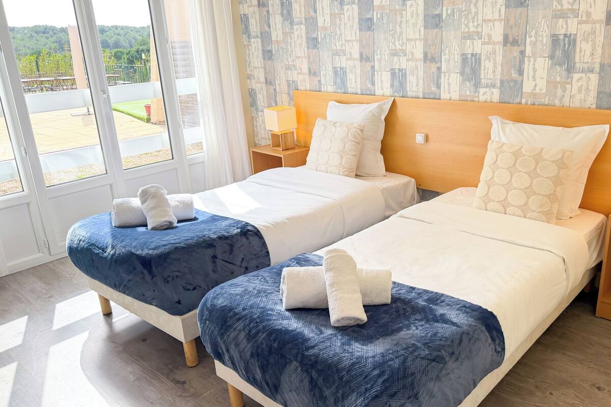 A hotel room with two beds, each with a blue bedspread and white linens, and a view of the outdoors through large windows.