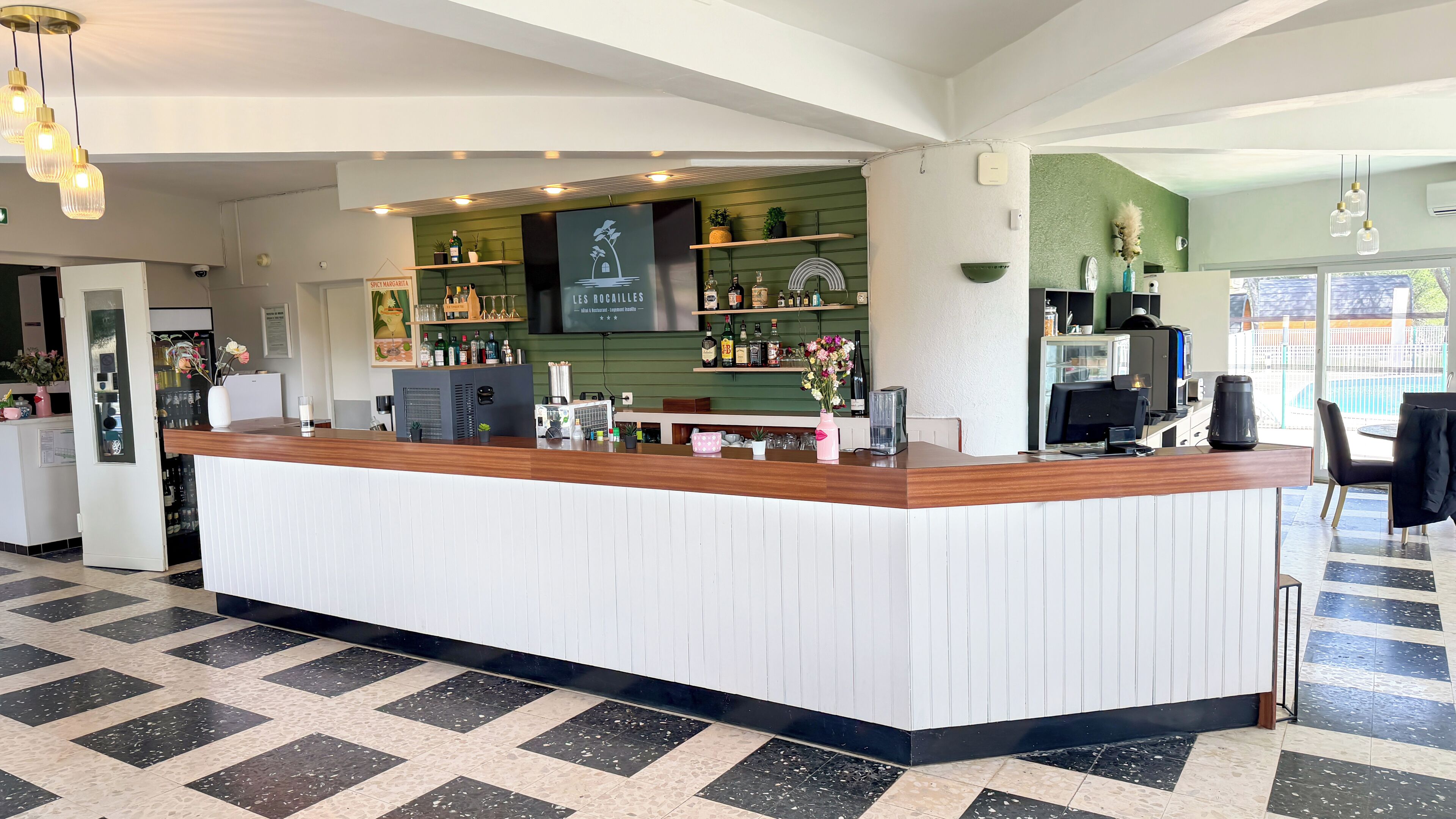 A modern reception area with a wooden counter, a green wall with shelves displaying bottles and glasses, and a large window providing natural light.