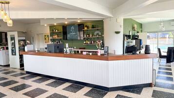 Un espace d’accueil moderne avec un comptoir en bois, un mur végétal agrémenté d’étagères où sont exposées des bouteilles et des verres, et une grande fenêtre qui laisse entrer la lumière naturelle.