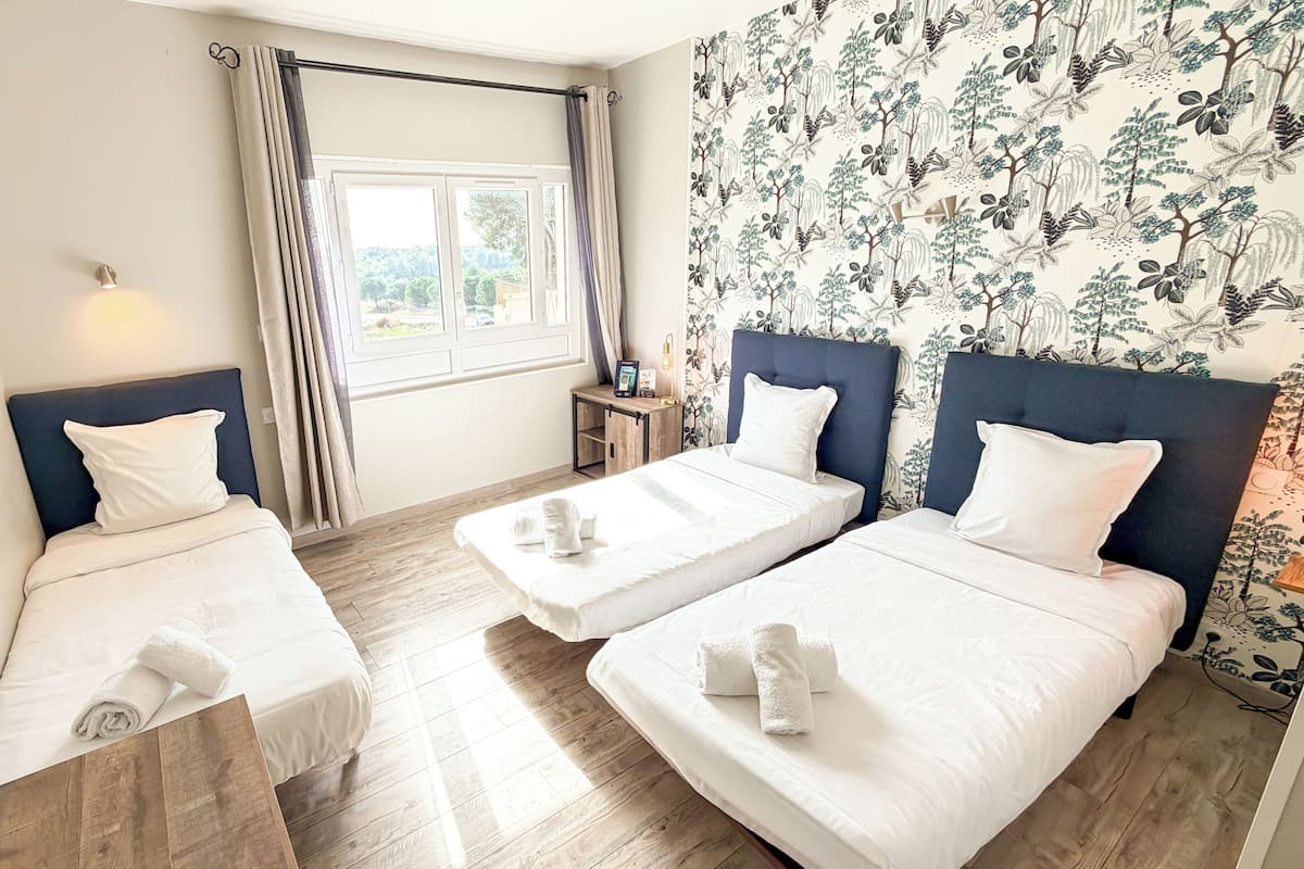 A hotel room with two single beds, a small bedside table with a lamp, a window with curtains, and a floral wallpaper.