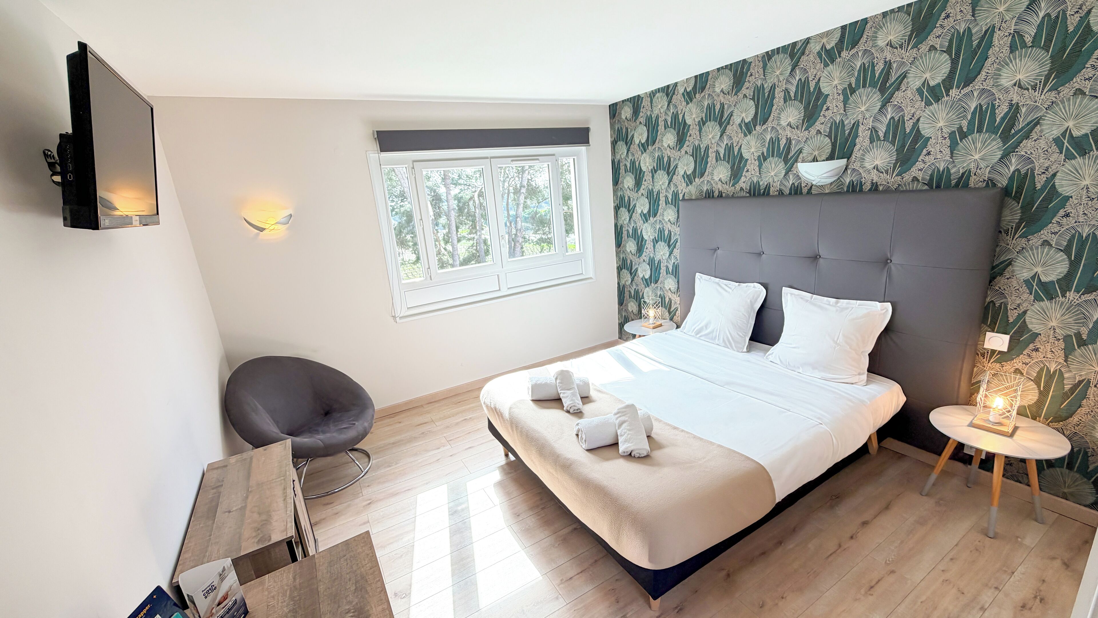A modern hotel room with a large bed, a grey chair, a small round table with a lamp, and a window with a view of trees.