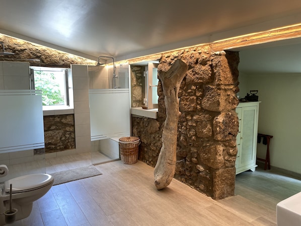 Une salle de bain avec un mur en pierre, un tronc d’arbre en bois, un meuble blanc et une fenêtre.