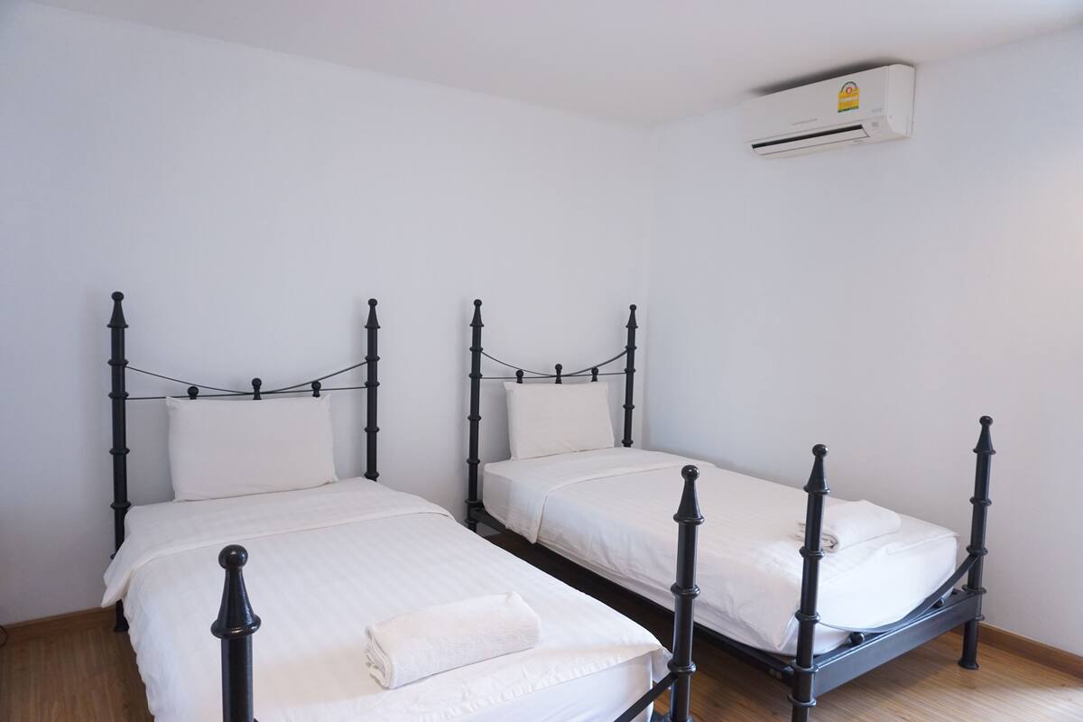 A room with three single beds, each with a black metal frame and white bedding. An air conditioning unit is mounted on the wall above the beds.