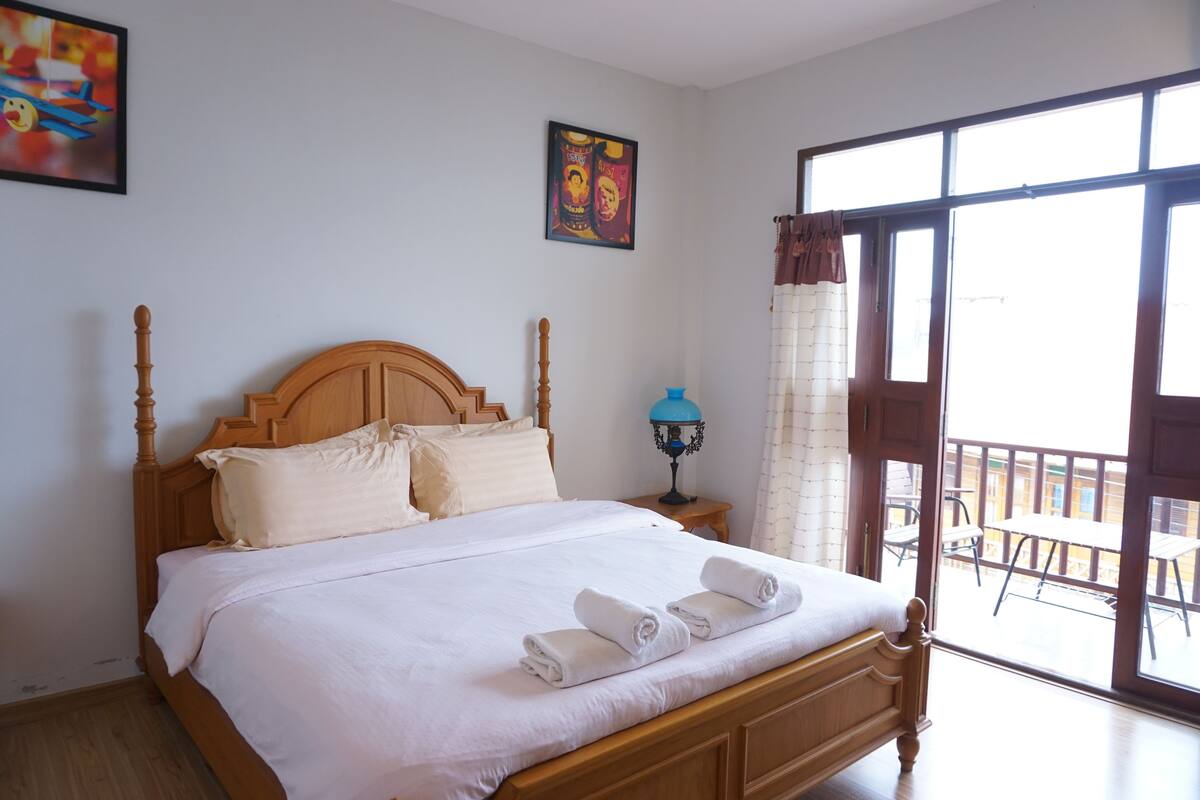 A neatly made bed with white linens and towels, a wooden headboard, a nightstand with a lamp, and a balcony with outdoor furniture.