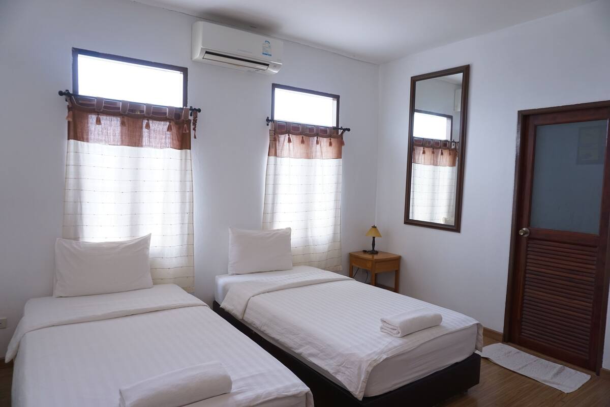 A room with two beds, a wooden door, a small table with a lamp, and a window with curtains.