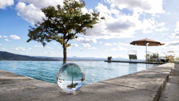 Una esfera de vidrio transparente que refleja un árbol y un entorno junto a una piscina con borde de hormigón, una silla y una sombrilla.