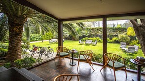 Varanda espaçosa com piso de madeira, cadeiras de vime e vista de um jardim bem cuidado, com gramado, palmeiras e uma bicicleta.