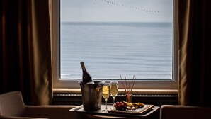 Quarto com vista para o mar e pássaros voando. Há uma mesa com uma garrafa, copos e comida.