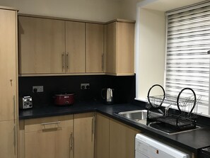 A kitchen with wooden cabinets, a black countertop, a red slow cooker, a kettle, and a dish rack.