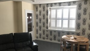 A living room with a black leather sofa, a wooden dining table with woven placemats, and a window with blinds. - Geise Farm Services (Thurso)
