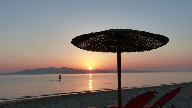 A beach scene with a sunset, a beach umbrella, and two red lounge chairs. - Evriali Luxury on the beach (Plaka Naxos)