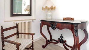 A room with a wooden chair, a table with a lamp, and a mirror on the wall. - [Le Grand Bleu] - Villa Maremonti Taormina (Taormina)