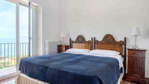 A bedroom with a large bed, a wooden headboard, two bedside tables with lamps, a radiator, and a balcony with a view. - [Le Grand Bleu] - Villa Maremonti Taormina (Taormina)