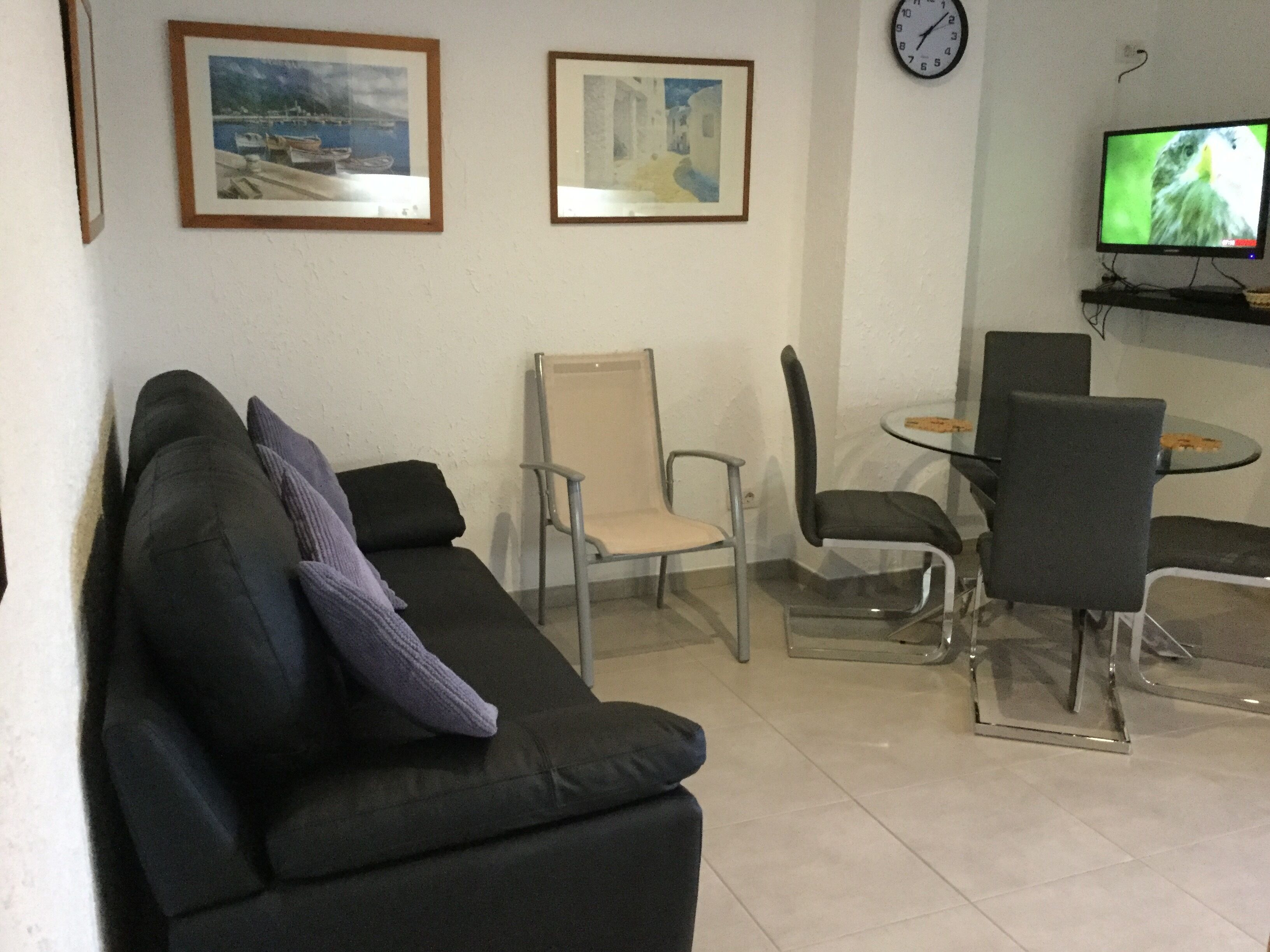 Una sala de estar con un sofá negro, dos sillones, una mesita y un televisor sobre un mueble.