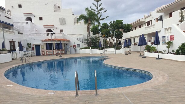A swimming pool with a tiled deck, surrounded by white buildings with balconies and blue umbrellas.