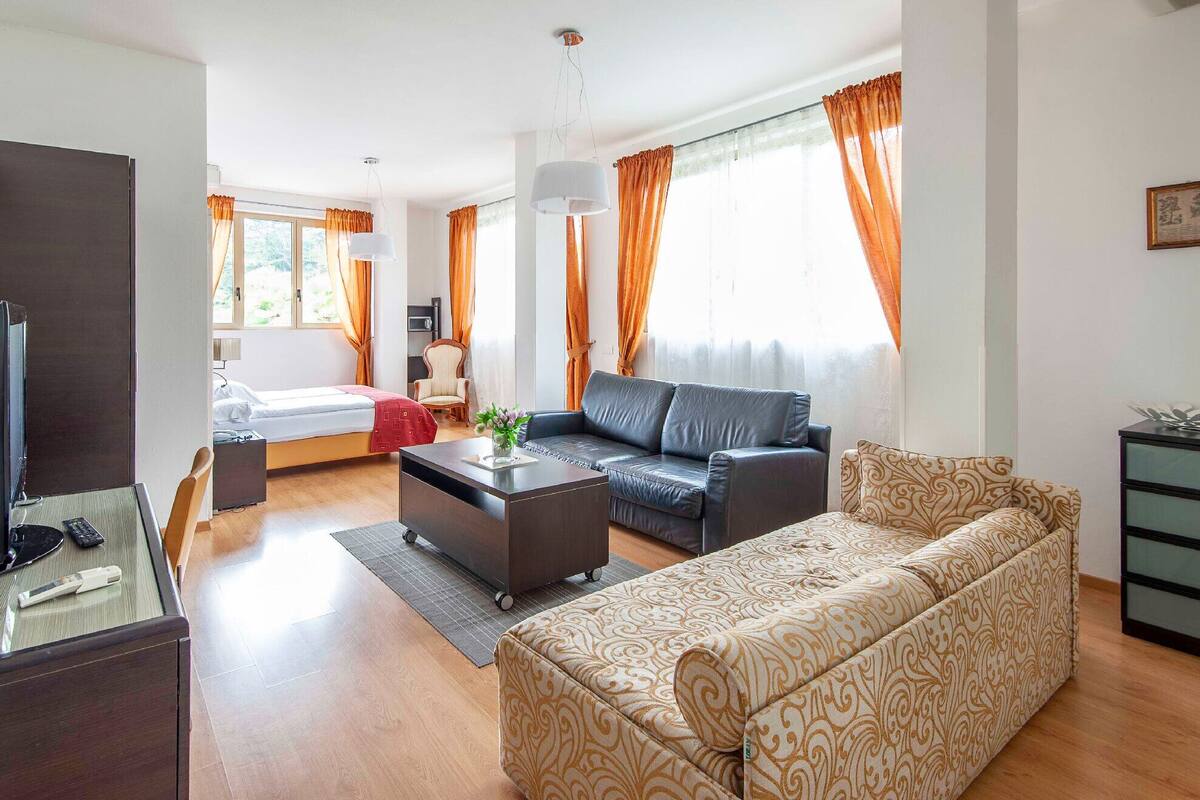 Un soggiorno moderno con divano, tavolino, televisione e un letto sullo sfondo.