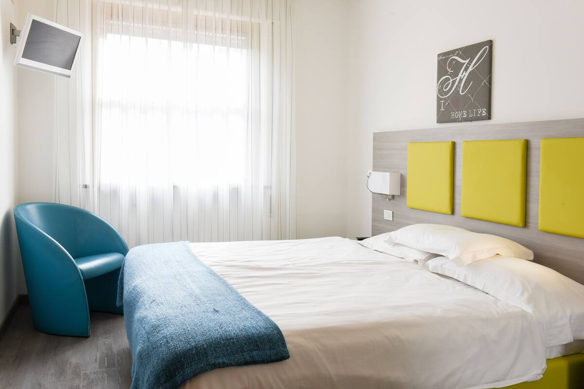 Una camera d'albergo con un letto, una poltrona blu, una televisione e un muro con un cartello.