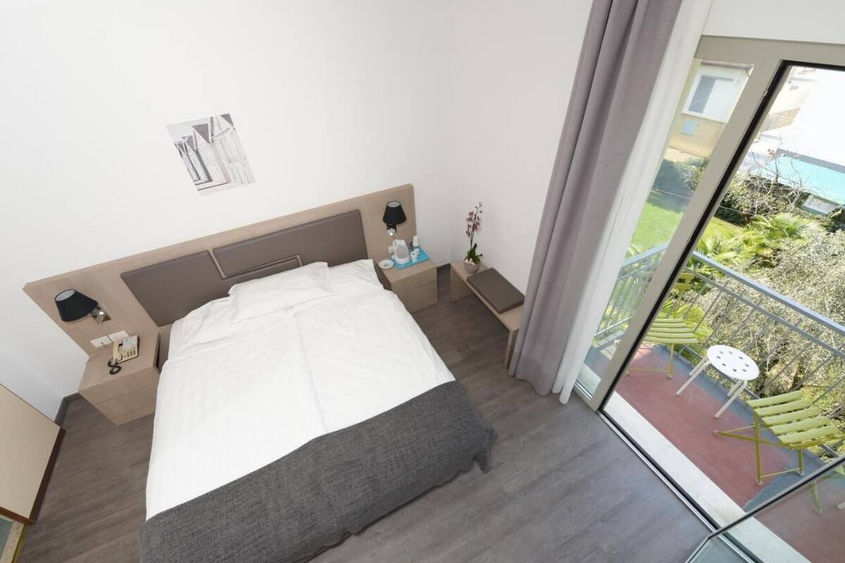 Una camera d'albergo moderna con un letto grande, un comodino con una lampada, un tavolino con una pianta e un balcone con mobili da esterno.